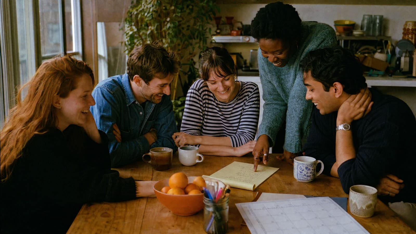 Five friends around a kitchen table planning a dinner party on a legal pad