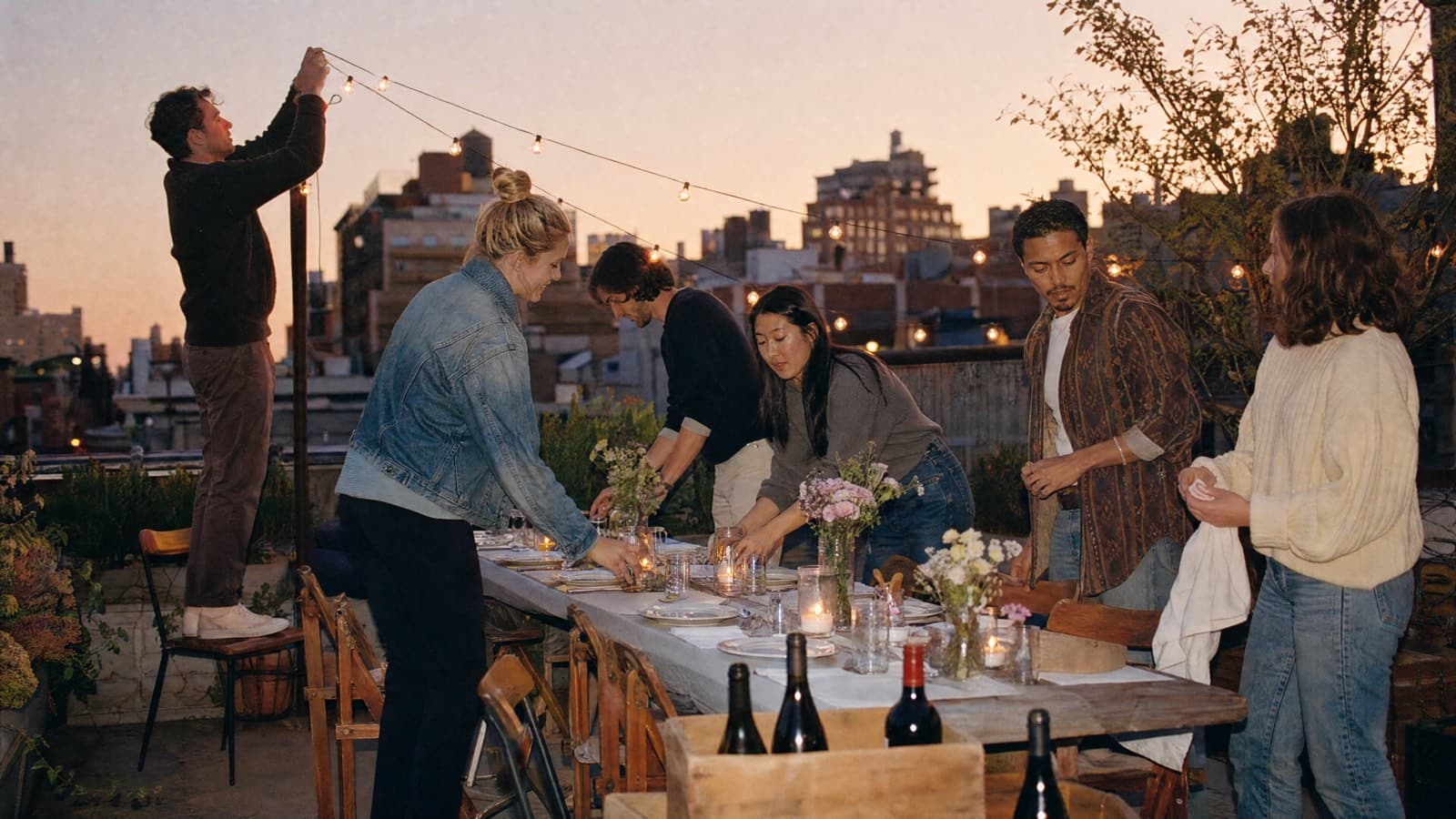 Friends setting up a long dinner table on a rooftop at golden hour, stringing lights and laying out candles