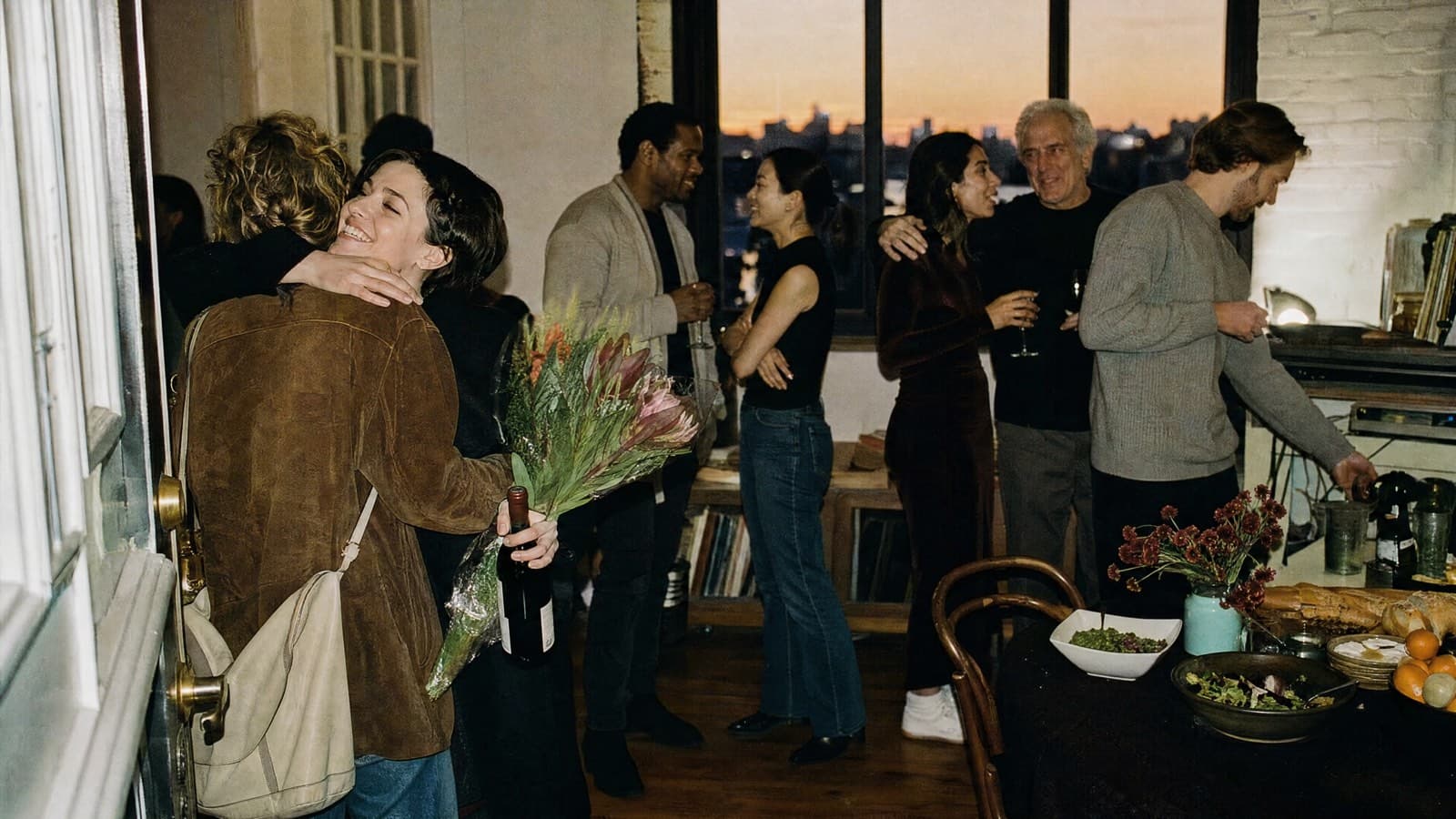 A host opening the door of a loft party and hugging a friend who arrives with wine and flowers